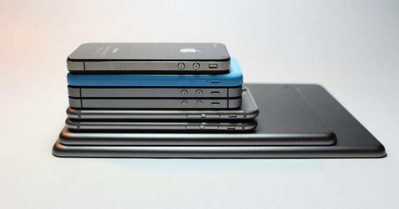 A stack of different smartphones. Starting with a tablet at the bottom, and going smaller to the top. In total there are 8 phones. The background is grey and blue-ish.