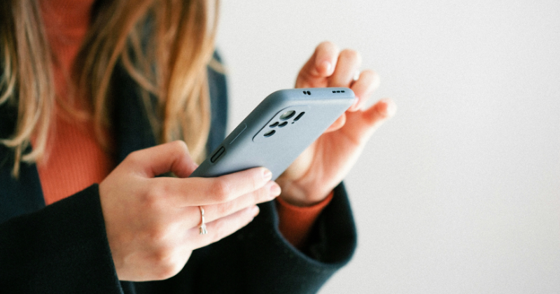 Image of a woman holding a blue iPhone in front of her. We only see her hands and her hair as she is holding the phone with two hands. There is a plain blue background.