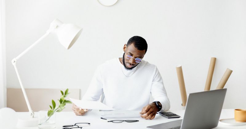 Man in a white sweater working at a desk with papers and a laptop.